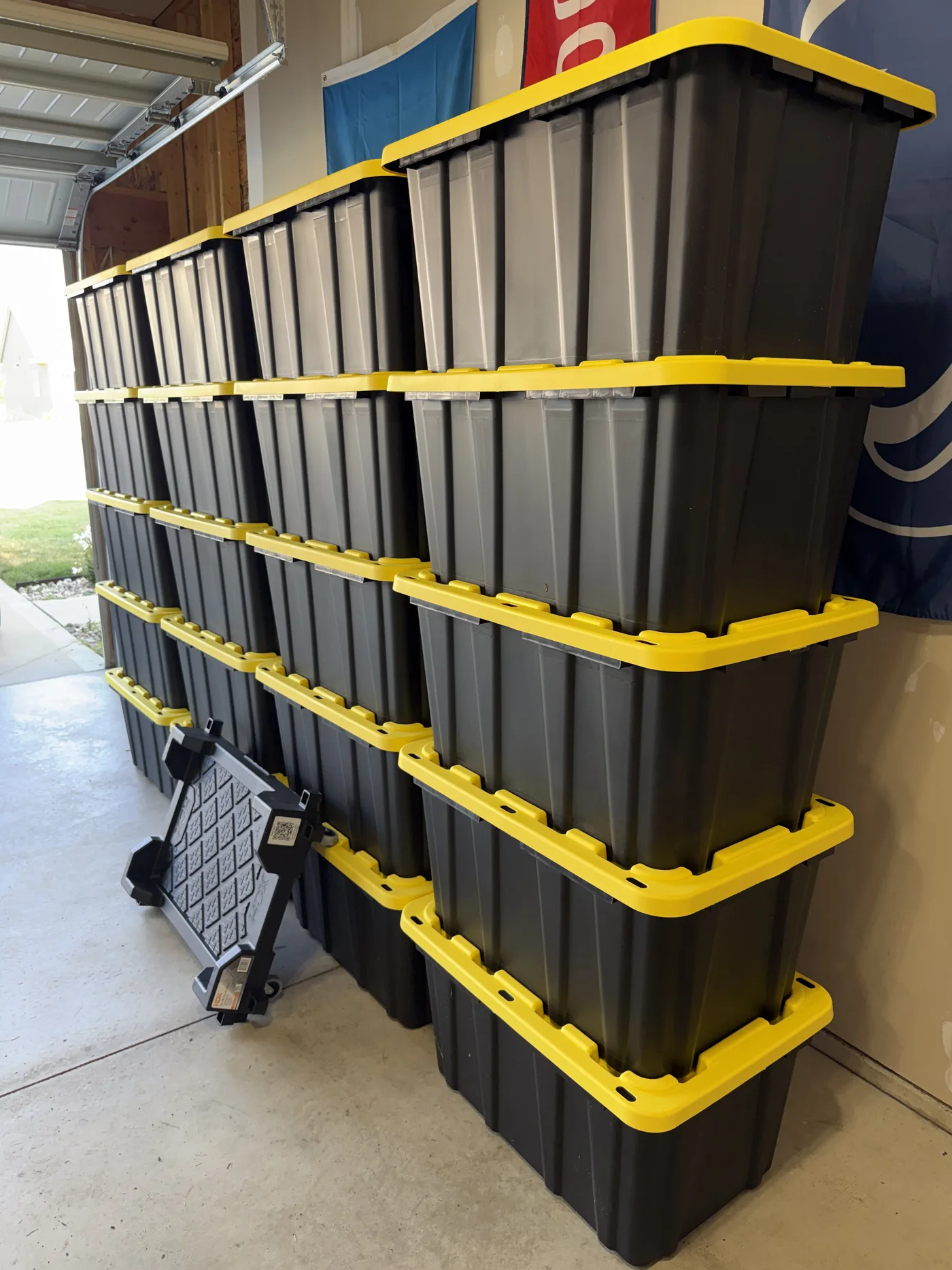 A tall, orderly stack of closed black moving totes with yellow lids